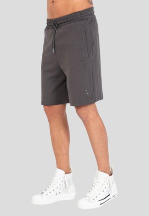 Hombre vestido con pantalones cortos gris oscuro hasta la rodilla con cintura elástica y zapatillas blancas de caña alta, de pie contra un fondo claro.
