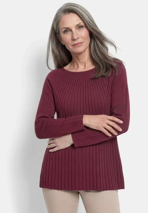 Reife Frau mit grauen Haaren, die einen gerippten, bordeauxfarbenen Langarmpullover und hellbeige Hosen trägt, steht mit verschränkten Armen.