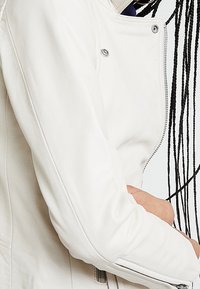 Bílá kožená motocyklová bunda s přiléhavým střihem, diagonálním zipem, stříbrným kováním a hladkou texturou.