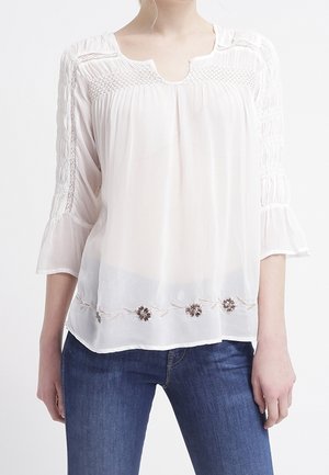 Femme portant un chemisier blanc à manches longues avec broderie florale à l'ourlet et un jean bleu en denim, debout, les bras détendus.