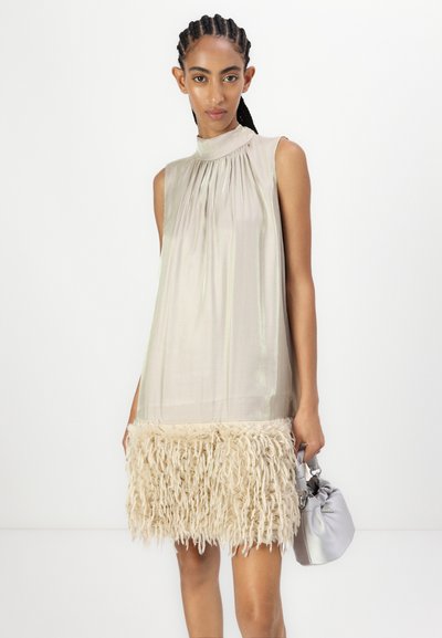 Robe sans manches avec encolure froncée en tissu beige clair, ornée d'une bordure en franges beige duveteuses à l'ourlet. Accentuée d'un sac gris.