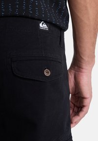 Černé šortky vyrobené z bavlny, s zadní kapsou opatřenou hnědým knoflíkem a štítkem s logem blízko pasu. Hladká textura, ležérní design.