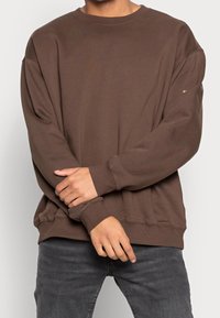 Felpa marrone in cotone con vestibilità rilassata, collo rotondo e maniche lunghe. Polsini e orlo a costine. Indossata con jeans grigi.