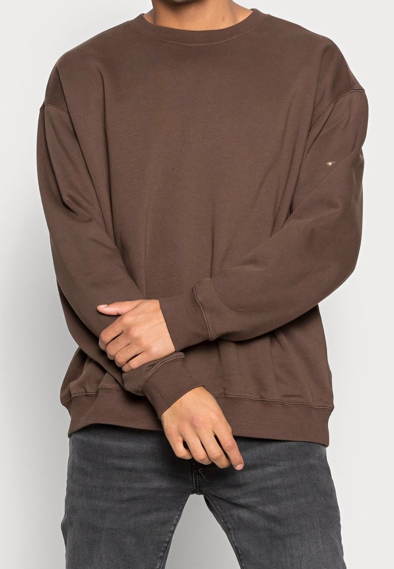 Felpa marrone in cotone con vestibilità rilassata, collo rotondo e maniche lunghe. Polsini e orlo a costine. Indossata con jeans grigi.