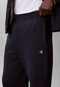 Uomo che indossa pantaloni della tuta Calvin Klein blu navy con vita elastica, t-shirt bianca e giacca nera, mano infilata vicino alla vita.