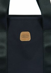 Borsa tote in tessuto blu navy con manici neri e un'etichetta in pelle marrone. Texture liscia e forma rettangolare con dettagli di cucitura rinforzata.