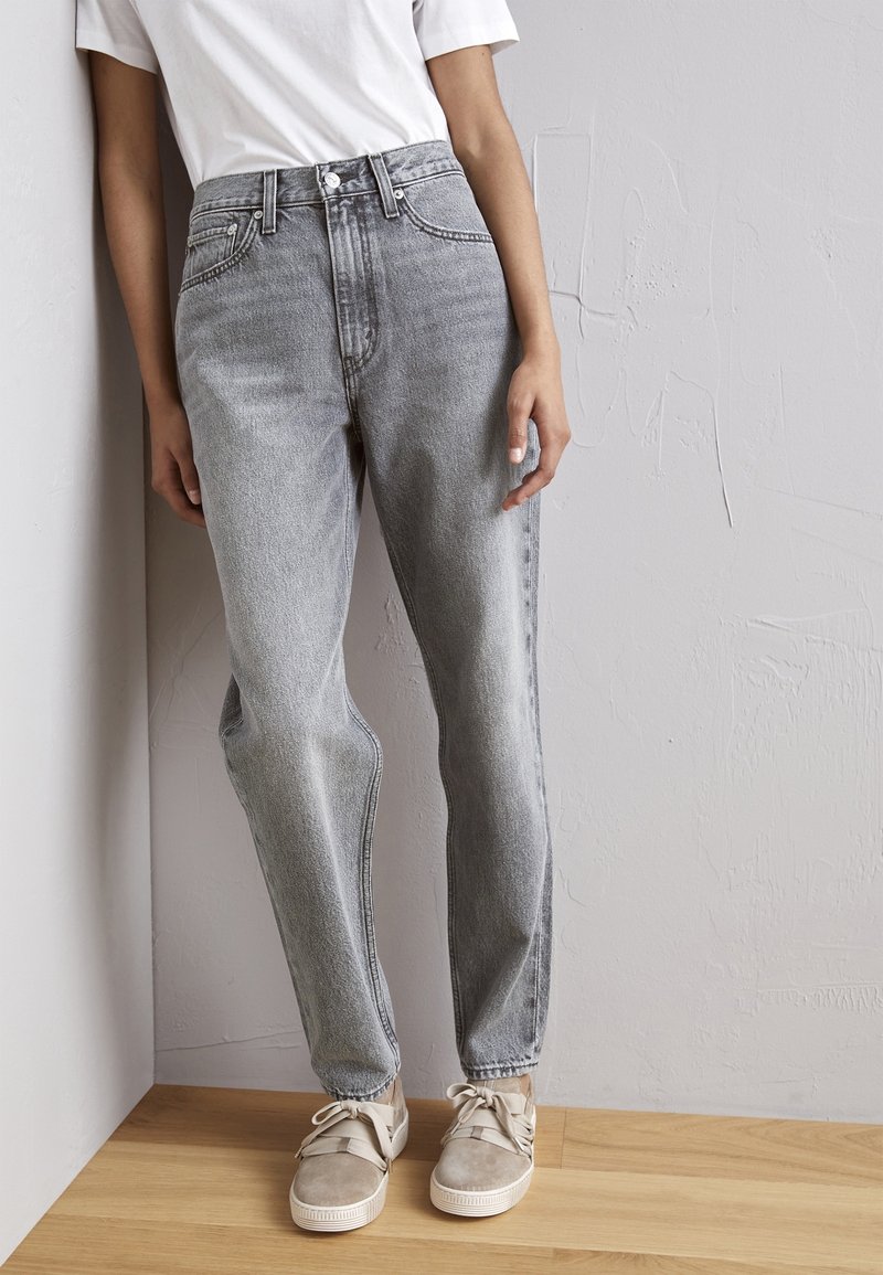Personne portant un jean gris taille haute, un t-shirt blanc et des baskets beiges, debout sur un sol en bois contre un mur gris clair.