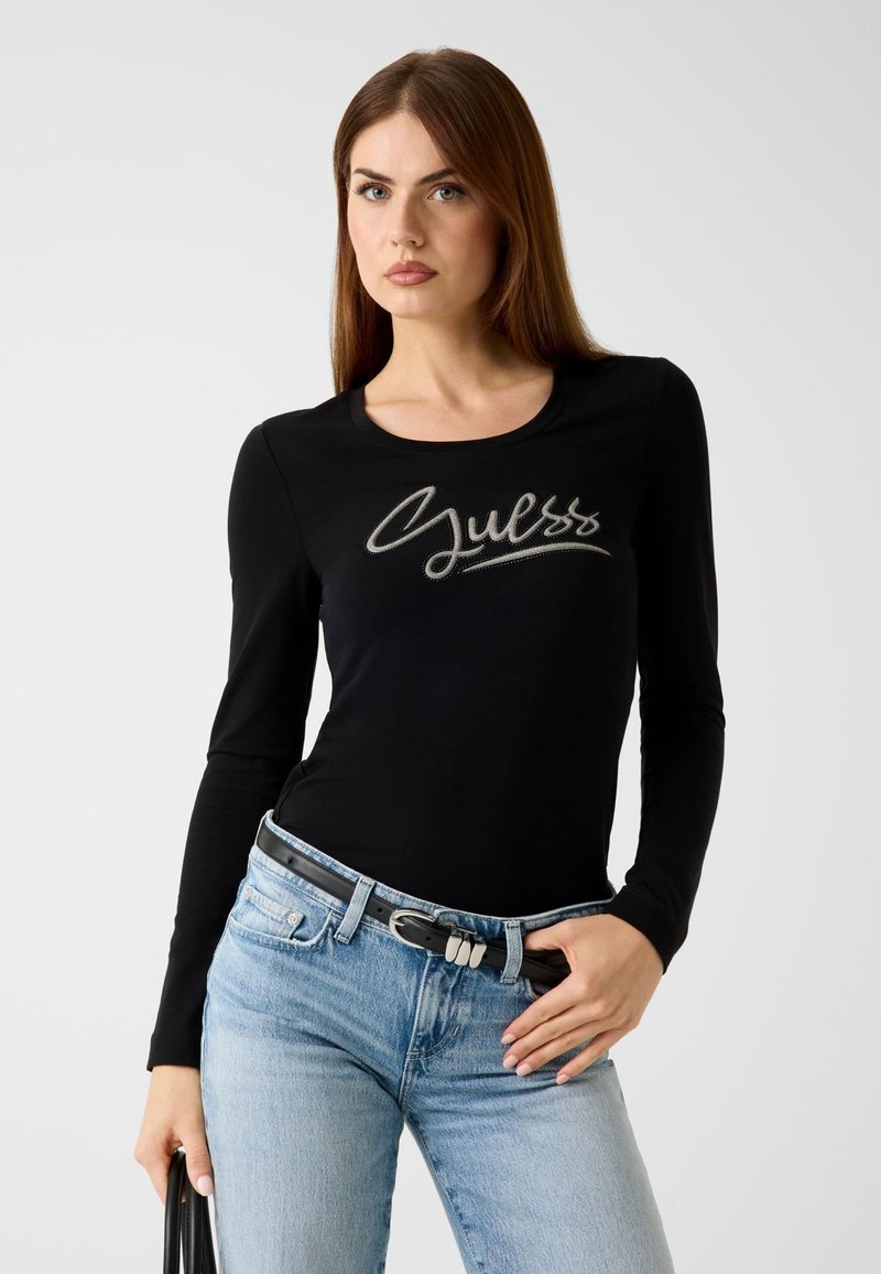 Schwarzes Langarmshirt mit rundem Ausschnitt und metallicfarbenem "Guess"-Logo auf der Vorderseite. Kombiniert mit hellblauen Jeans und einem schwarzen Gürtel.
