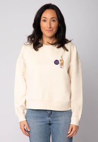 Sweatshirt couleur crème à coupe décontractée, avec un petit graphique d'un chat et du texte sur le devant. Matière en mélange de coton doux.