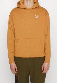 Felpa arancione con tasca frontale, polsini a costine e un piccolo logo Puma bianco sul petto. Il tessuto sembra morbido e strutturato.