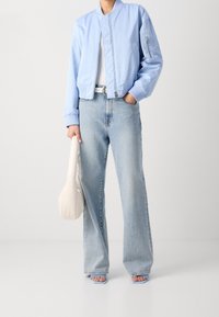 Jasnoniebieska kurtka bomber z prążkowanymi mankietami i kołnierzem, w zestawieniu z jasnymi, wysokim stanem jeansami. Trzyma kremową saszetkę.