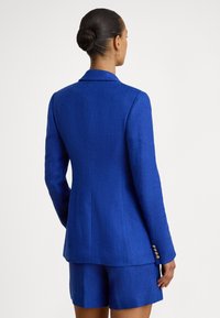 Modri blazer z ribjo kostjo in krojenim krojem, z enojnim zapenjanjem na gumb in tremi gumbi na manšetah. Oblečen z usklajenimi modrimi kratkimi hlačami.