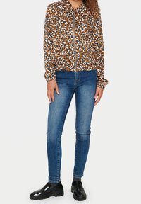 Kvinna klädd i en brun, marinblå och vit leopardmönstrad zip-jacka, blå skinny jeans, svarta loafers och grå strumpor, stående mot vit bakgrund.