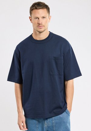 Homme aux cheveux courts portant un T-shirt oversize bleu marine avec poche et un jean bleu clair, debout avec une main dans la poche.