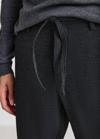 Schwarze, strukturierte Hose mit elastischem Bund und Kordelzug. Das Material hat einen dezenten Glanz und zeigt ein feines Muster durchgehend.