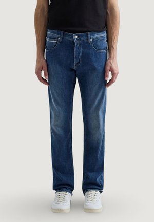 Hombre con vaqueros de corte recto de mezclilla azul y zapatillas blancas, de pie frente a un fondo claro y liso.