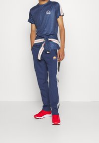 Granatowa koszulka sportowa z logotypem, z ciemnoniebieską, wzorzystą tkaniną i paskami na rękawach. Dopasowane granatowe spodnie z bocznymi paskami i czerwone sneakersy.