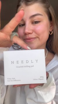 Un emballage rectangulaire blanc étiqueté "NEEDLY Cicachid Chilling Pad." Texte en gris. Présenté dans une main, mettant en avant la surface lisse et le design minimaliste.