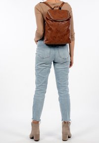 En person som bär ljusblå jeans, beige klackboots och en brun läderryggsäck, står mot en vit bakgrund.