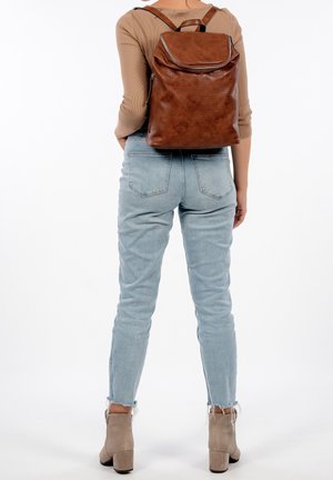 Person in hellblauen Jeans, beigen Absatzstiefeln und einem braunen Leder-Rucksack, der vor einem weißen Hintergrund steht.