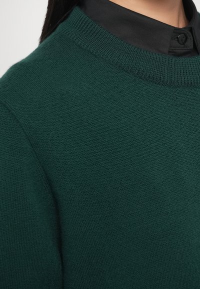 Tumši zaļš adīts džemperis ar apaļu izgriezumu un mīkstu, teksturētu audumu, nēsāts virs melnas apkaklītes krekla.