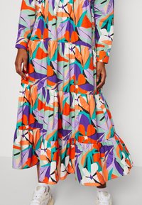 Robe maxi florale colorée avec de longs manches, présentant des étagères de volants, dans des teintes d'orange, de violet et de vert sur un fond clair.