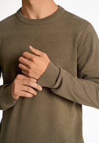 Maglione verde oliva con strisce orizzontali testurizzate, dotato di scollatura e polsini a costine. Materiale lavorato morbido, design aderente.