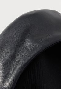 Musta nahkahattu, jossa on teksturoitu pinta ja ommeltu vuori sisäpuolella. Materiaaliin painettu hienovarainen brändäys.