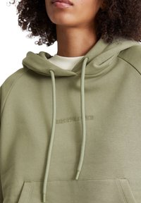 Olivgrön hoodie med dragsko i huvan, ribbade ärmslut och subtilt broderad logotyp framtill. Slät, mjuk tygkvalitet med avslappnad passform.