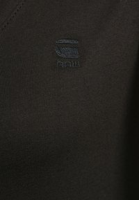 Svart bomullsplagg med mjuk textur, med en subtil logotyp broderad i mörkblått. Designen inkluderar en v-ringad hals och ribbad tyg.