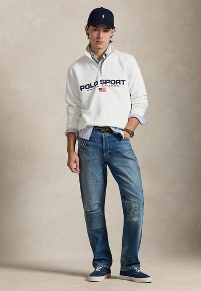 Balts vilnas džemperis ar "POLO SPORT" logotipu, zilas džinsa bikses ar nodilušām vietām un tumši zili sporta apavi, valkāti ar cepuri un slāņotu kreklu.