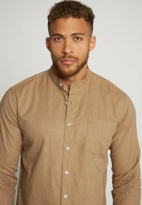 Camisa marrón de manga larga con cuello tipo banda, bolsillo frontal y cierre de botones. La tela tiene una apariencia tejida y texturizada.