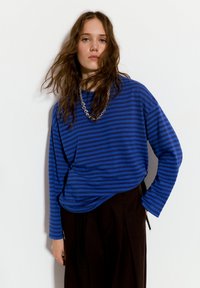 Mladá žena s dlouhými vlnitými hnědými vlasy, oblečená v modro-černém pruhovaném tričku s dlouhým rukávem, černých kalhotách a stříbrném řetízku.