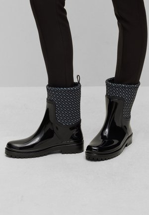 Bottes de pluie noires brillantes à la cheville avec des poignets en tissu à motifs portées avec un pantalon noir slim sur un sol gris clair.