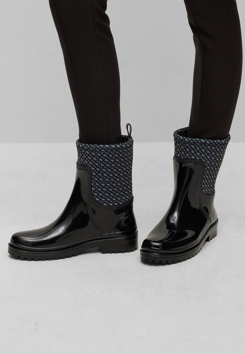 Bottes de pluie noires brillantes à la cheville avec des poignets en tissu à motifs portées avec un pantalon noir slim sur un sol gris clair.