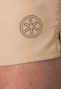 Šviesiai kreminiai šortai, pagaminti iš lygaus audinio, su apvaliu, išsiuvinėtu emblema, turinčia žvaigždės formą, ir siuvinėta apvadu.