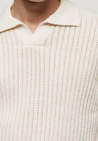 Polo de punto color crema con cuello en V, que cuenta con rayas verticales y un patrón texturizado en toda la tela.