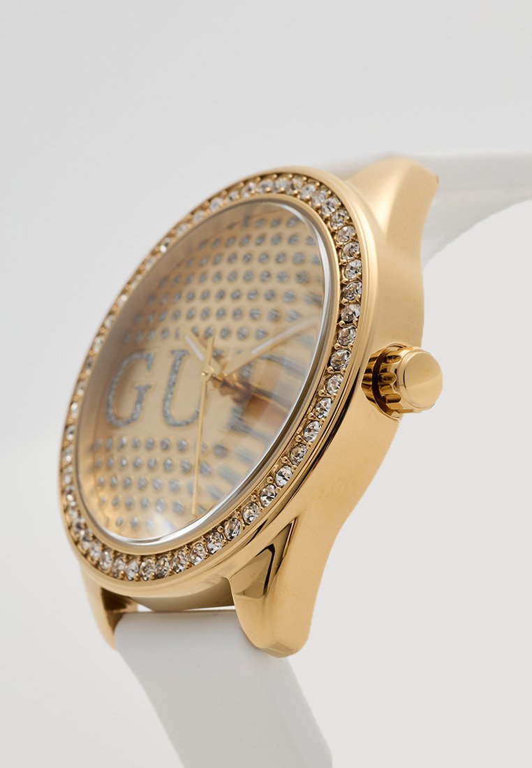 Reloj de tono dorado con una esfera transparente que presenta un patrón con tachuelas, adornado con un bisel de cristal y una correa de silicona blanca.