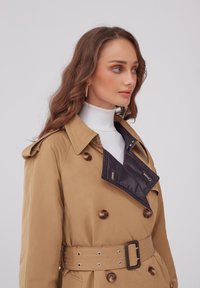 Beżowy trencz z podwójnymi guzikami, oversized klapami i paskiem w talii, z brązowymi guzikami i kontrastującym wewnętrznym kołnierzem.