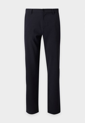 Homme debout portant un t-shirt blanc uni à manches courtes, un pantalon noir et des baskets blanches sur fond blanc.