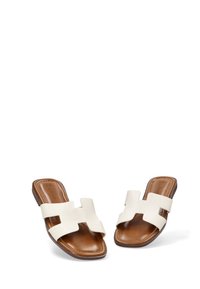 Chanclas planas con un empeine sintético blanco que presenta un diseño de tira ancha, montadas sobre una suela marrón similar al cuero. Textura suave en general.