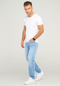 White short-sleeve t-shirt, light blue jeans, and white sneakers. Model is standing with one hand in pocket against a plain background.