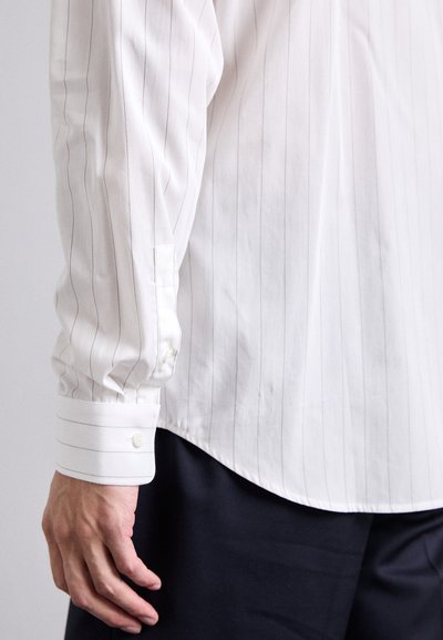Camisa blanca de manga larga con finas rayas verticales, con puños abotonados y textura suave, combinada con pantalones oscuros.