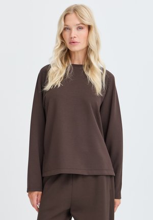 Blonde vrouw met een losse donkerbruine top met lange mouwen en bijpassende broek, staande tegen een effen lichte achtergrond.