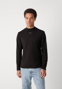 Must mustaga, kõrge kaelusega must särk pika varrukaga, millel on rindkeres valge "Calvin Klein" logo, valmistatud siledast ja venivast puuvillaste materjalidest.