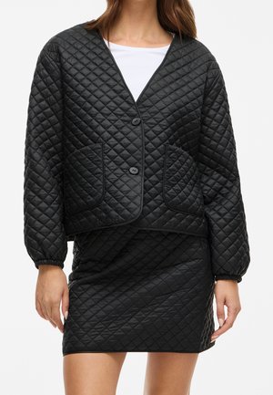 Femme portant une veste matelassée noire avec des boutons et une jupe matelassée assortie, associées à un haut blanc en dessous.