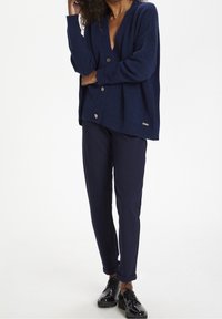 Mörkblå cardigan med djup V-ringning, strukturerat tyg och silverknappar, ihopparad med mörkblå byxor och blanka svarta skor.