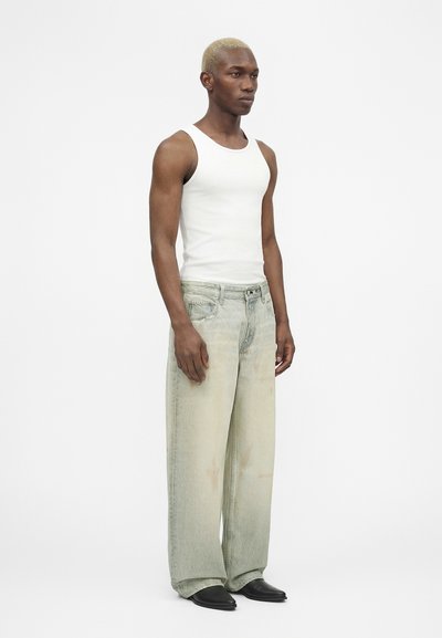 Άνδρας με κοντά ξανθά μαλλιά που φοράει λευκό αμάνικο τοπ, ξεθωριασμένο τζιν με φαρδιά μπατζάκια και μαύρα μυτερά παπούτσια, στέκεται μπροστά σε απλό φόντο.