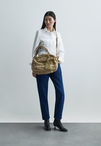 Miękka beżowa skórzana torba z podwójnymi uchwytami i długim paskiem, z detalami przeszyć i metką bagażową, noszona z białą koszulą i niebieskimi dżinsami.