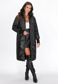 Sort polstret jakke med høj krave, fuldlængde design og ribbede manchetter; har lynlåslukning og blank overflade. Bæres over en hvid top.